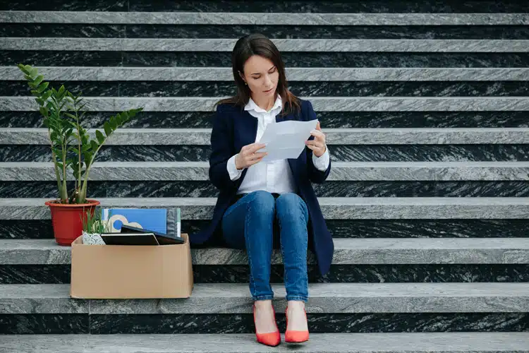 A woman sitting on stairs reviewing a letter, surrounded by a box of office items and a potted plant.