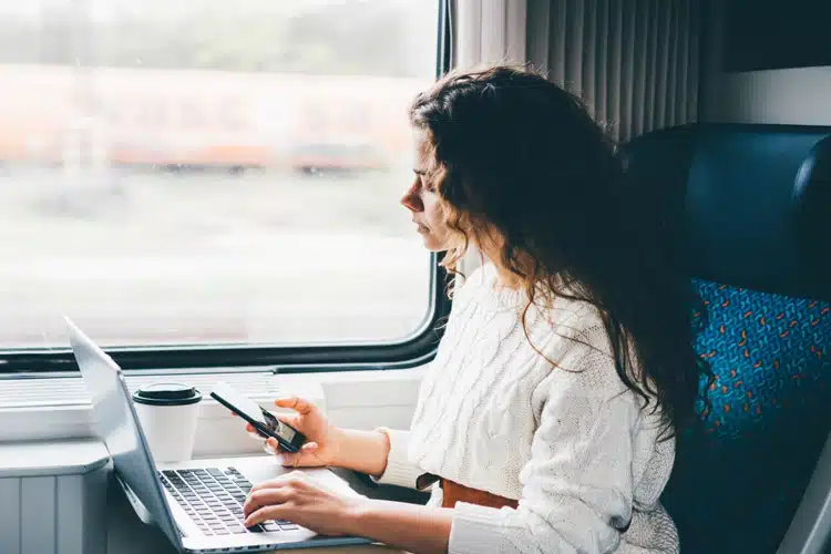 woman sitting in a train looking at her phone with a laptop in-front of her